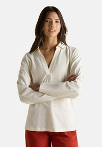 Pikkade tumedate juustega naine, kes kannab kreemivärvi pika varrukaga V-kaelusega särki ja punaseid pükse, seisab käed ristis ühtlasel taustal.