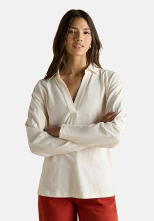 Žena s dlhými tmavými vlasmi, oblečená v krémovom tričku s dlhým rukávom a výstrihom do V, a červených nohaviciach, stojí so založenými rukami pred jednoduchým pozadím.