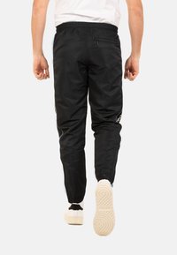Pantalons de jogging noirs avec taille élastique, jambes fuselées, zippers latéraux et un logo blanc discret sur la hanche gauche. Les chaussures visibles sont blanches avec des accents noirs.