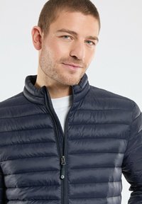 Veste matelassée bleu marine avec un col montant et une fermeture éclair à l'avant. Présente un motif matelassé horizontal et une texture lisse et brillante.