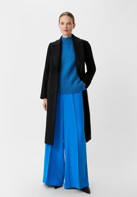 Schwarzer langer Mantel, leuchtend blauer strukturierter Pullover und weit geschnittene blaue Hose. Das Ensemble bietet eine moderne Passform mit klaren Linien.