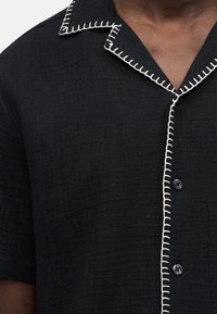 Chemise noire texturée à manches courtes avec bordure blanche cousue délimitant le col et la patte de boutonnage, portée par une personne à la peau foncée.