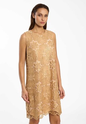 Frau trägt ein ärmelloses, beiges Spitzenkleid mit Blumenmuster und steht vor einem schlichten weißen Hintergrund.