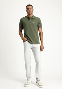 Ciemnozielona koszulka polo z klasycznym kołnierzykiem, krótkimi rękawami i małym logo; zestawiona z jasnoszarymi jeansami o slim-fit i białymi butami sportowymi.