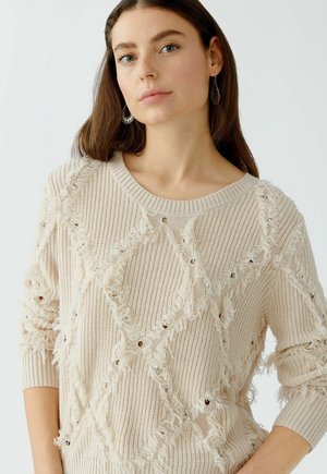 Beige gebreide trui met een textuurpatroon, voorzien van rafelige accenten en metalen oogjes, ontworpen met een ronde halslijn en lange mouwen.