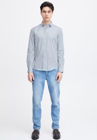 Lichtblauw overhemd met lange mouwen en een puntige kraag, gecombineerd met faded blauwe jeans en zwarte schoenen, gepresenteerd van voren tegen een witte achtergrond.