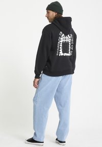 Un sweat à capuche noir avec un imprimé graphique sur le dos, associé à un jean baggy bleu clair. Personne portant un bonnet vert et des baskets noires.