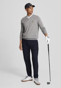 Maglione grigio a collo a V con logo giallo, abbinato a pantaloni blu. Il modello tiene un bastone da golf, indossando un berretto blu e guanti, con scarpe da ginnastica bianche.