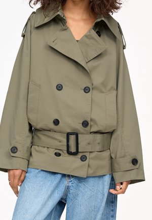 Trench-coat croisé vert olive avec boutons noirs et ceinture, porté sur un jean bleu clair, visible du cou aux hanches.