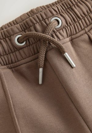 Brūnas sporta bikses ar elastīgu jostasvietu, sudraba acīm un pinuma auklu ar metāla galiņiem. Mīksts, teksturēts audums.