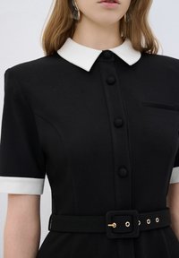 Schwarzes Kleid mit weißem Kragen und Manschetten, kurzen Ärmeln, Knopfleiste und einem Gürtel mit einer goldenen Schnalle. Verfügt über eine Brusttasche.