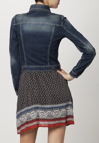 Mörkblå denimjacka med figurnära passform, klassiska sömmar och knappdetaljer. Nedanför en mönstrad klänning med spetskant.