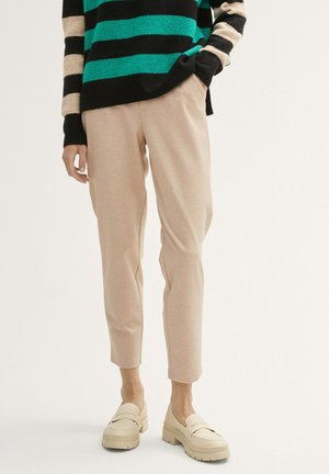Pantalon beige fuselé avec poches latérales, associé à un pull rayé noir et vert. Modèle portant des mocassins crème.