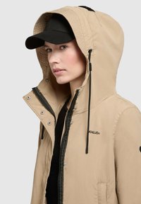Chaqueta con capucha de color beige claro, con cremallera frontal negra y detalles en negro, que incluye botones a presión y cordones. Una gorra negra completa el conjunto.