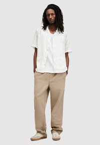Hombre con dreadlocks largos que lleva pantalones beige holgados, zapatillas blancas, una camisa blanca de manga corta texturizada y una camiseta interior blanca de canalé, de pie.