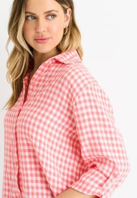 Camicia a righe gingham rosa e bianca con colletto e maniche arrotolate. Il tessuto sembra leggero e ha una texture liscia.