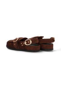Zapatos de ante marrón con suela plana, que presentan dos correas anchas con hebillas doradas y costuras decorativas en la zona de los dedos.