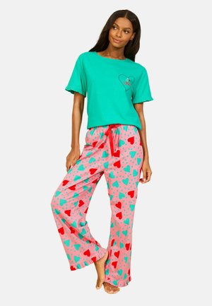 Femeie care poartă un tricou verde cu mâneci scurte și pantaloni de pijama roz cu modele de inimi roșii și turcoaz, stând desculță cu un picior ușor ridicat.