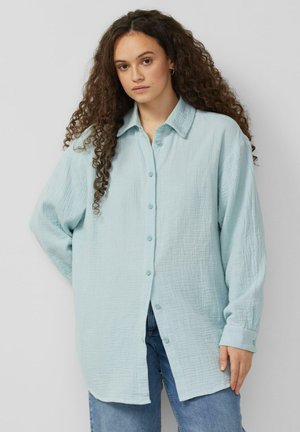 Femme aux longs cheveux bouclés portant une chemise ample bleu clair à boutons et un jean bleu, se tenant devant un fond uni.