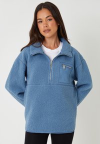 Blå fleecepullover med halv-zip lukning, en frontlomme i en kontrastfarve og en blød, tekstureret overflade.