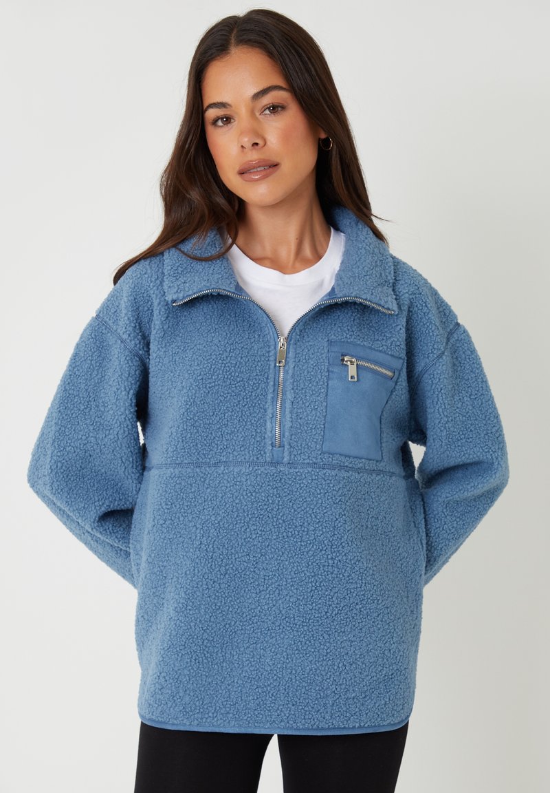 Blå fleecepullover med halv-zip lukning, en frontlomme i en kontrastfarve og en blød, tekstureret overflade.