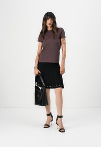 T-shirt marron à manches courtes avec logo en relief, associé à une jupe plissée noire avec des accents métalliques, et un sac à main en cuir noir.