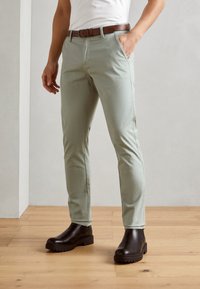 Lichtgroene, slim-fit broek van katoen, met zijzakken en een bruine riem. Gemaakt met bruine boots met dikke zolen op een houten vloer.