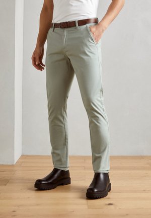 Lichtgroene, slim-fit broek van katoen, met zijzakken en een bruine riem. Gemaakt met bruine boots met dikke zolen op een houten vloer.