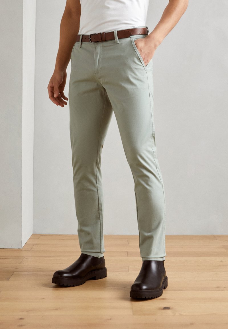 Lichtgroene, slim-fit broek van katoen, met zijzakken en een bruine riem. Gemaakt met bruine boots met dikke zolen op een houten vloer.