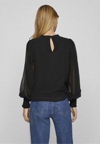 Blusa nera con maniche a sbuffo trasparenti, dettaglio a forma di chiave sul retro e tessuto del corpo in tinta unita. Abbinata a jeans di denim blu.
