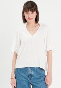 White blouse with a V-neck, short embroidered sleeves featuring eyelet details. Soft fabric with a textured dot pattern. Paired with blue jeans.