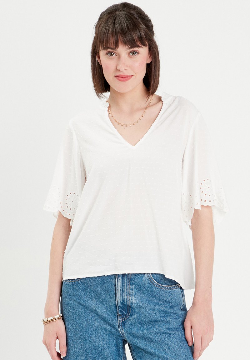 White blouse with a V-neck, short embroidered sleeves featuring eyelet details. Soft fabric with a textured dot pattern. Paired with blue jeans.