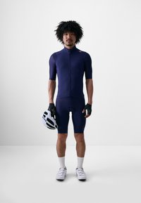 Navy cyklistický dres s krátkymi rukávmi a predným zipsom. Nosí sa s čiernymi rukavicami, bielymi cyklistickými topánkami a bielou prilbou. Hladká textúra tkaniny.