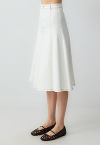 Weiße Denim-A-Linien-Rock mit hoher Taille, einem Reißverschluss auf der Vorderseite und sichtbaren Nähdetails; kombiniert mit braunen geflochtenen Ballerinas.