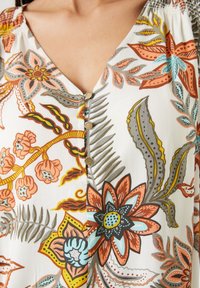 Blusa floral con escote en V, con un fondo blanco adornado con flores de colores en naranja, azul y verde. Incluye botones decorativos.