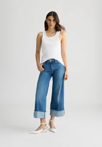 Valge ribaga tank-top logo'ga, koos helesiniste laiade denim-pükstega, mille alumine äär on volditud, ja valgete pahkluu-ribadega kontsadega.
