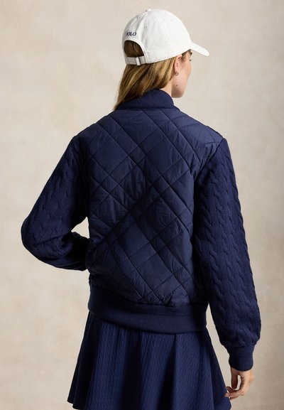 Femme portant une veste matelassée bleu marine avec des manches torsadées, une jupe bleu marine et une casquette blanche, debout, dos à la caméra.