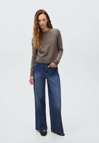 Hellbrauner Pullover mit gerippten Bündchen und dekorativen Knöpfen an der Schulter; kombiniert mit weiten, dunkelblauen Jeans.