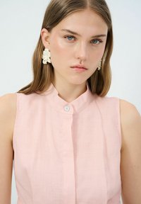 Blusa senza maniche rosa chiaro con colletto a listino, chiusura frontale con bottoni e delicati dettagli con bottoni. La modella indossa orecchini a forma di fiore.