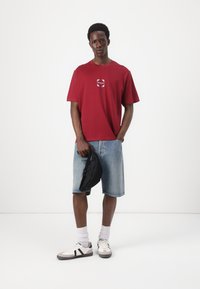 Red cotton t-shirt with a circular logo, paired with light blue denim shorts. Model wears black sneakers and holds a black bag.