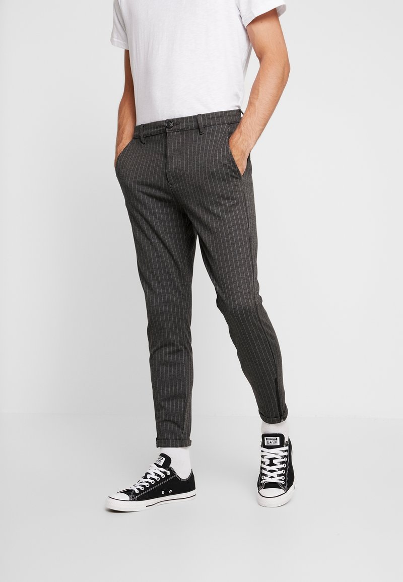 Szare spodnie w cienkie prążki o wąskim kroju i zwężanych nogawkach, wyposażone w boczne kieszenie i zapięcie na guzik, zestawione z czarnymi trampkami.