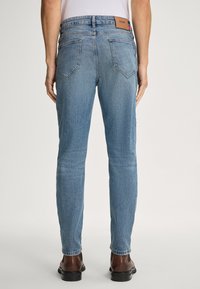 Mężczyzna ubrany w jasnoniebieskie dżinsy i brązowe skórzane buty, stojący plecami do widza na jednolitym tle.