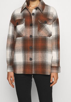 Persoon die een bruin-wit-zwart geruit overhemd met twee borstzakken en een zwarte broek draagt.
