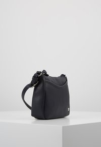 Geantă mică tip crossbody din piele neagră, cu finisaj texturat, curea reglabilă și logo metalic subtil, expusă pe o platformă albă.