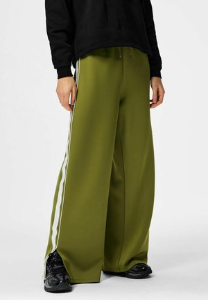 Person iført olivengrønne bukser med vide ben og hvide striber i siden, sorte sneakers og en sort langærmet top mod en hvid baggrund.