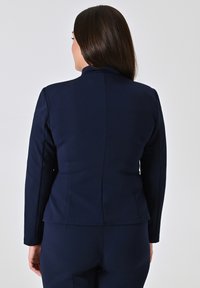Navyblauwe getailleerde blazer met een hoge kraag, een gladde textuur en gestikte details langs de achterkant en mouwen, gecombineerd met bijpassende broek.