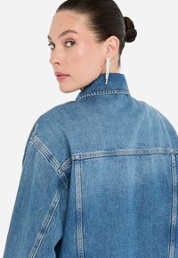 Giubbotto di jeans blu chiaro con colletto, cuciture visibili e vestibilità over. Presenta una yoke posteriore e orlo dritto. Orecchini a strati aggiuntivi.