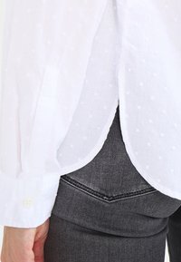 Chemise blanche avec un motif à pois texturé, manches longues, poignets à boutons et un ourlet arrondi, assortie à un jean en denim gris foncé.