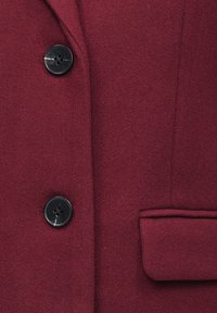 Λεπτομέρεια παλτού από μίγμα μαλλιού μπορντό, με μεγάλο μαύρο κουμπί, τσέπη με καπάκι και ομαλή υφή με λεπτό φινίρισμα και ραφές ραμμένες.