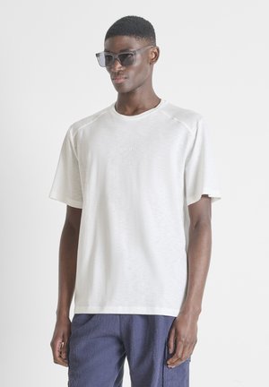 Giovane uomo con occhiali da sole grigi, t-shirt bianca a maniche corte e pantaloni blu con trama, in piedi su uno sfondo bianco uniforme.